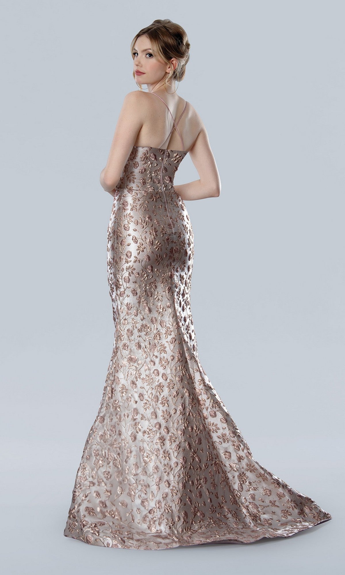 A woman in the Stella Couture 25328 metallic, floor-length formal dress with floral patterns stands looking over her shoulder against a plain background.